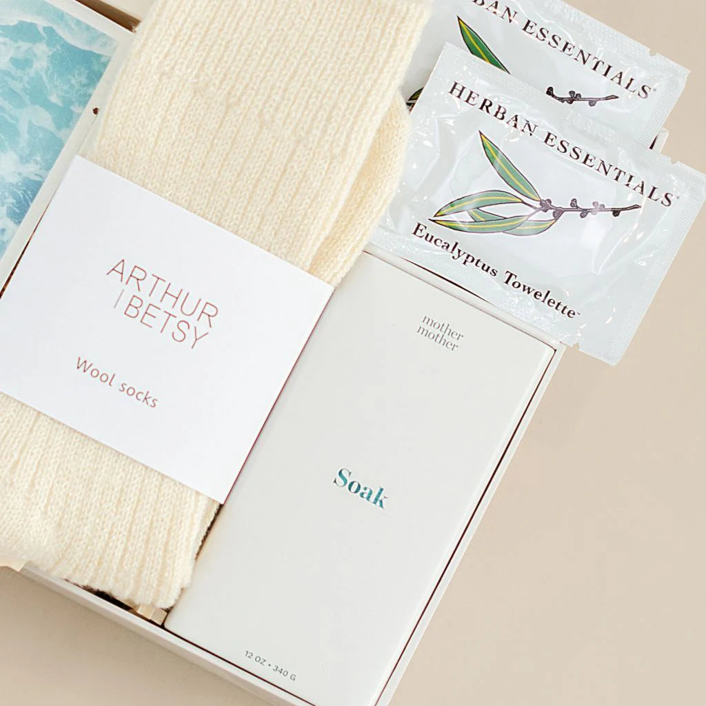 Product packaging with 'Arthur & Betsy' wool socks, 'Herban Essentials' towelettes, and 'Soak' product on a beige surface.