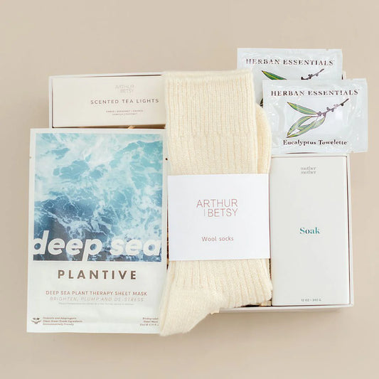 Collection of wellness products including a sheet mask, socks, tea lights, bath soak and eucalyptus towelettes on a beige background.