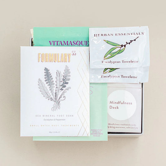 Collection of wellness products including a Vitamasque mask, Eucalyptus Towelette, Sea Mineral Foot Soap, and a Mindfulness Deck on a beige background.