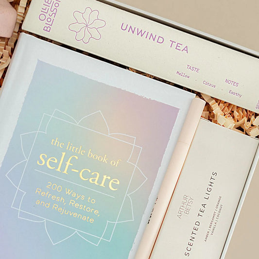 Book titled 'The Little Book of Self-Care' with a box of 'Unwind Tea' and 'Scented Tea Lights' in the background.